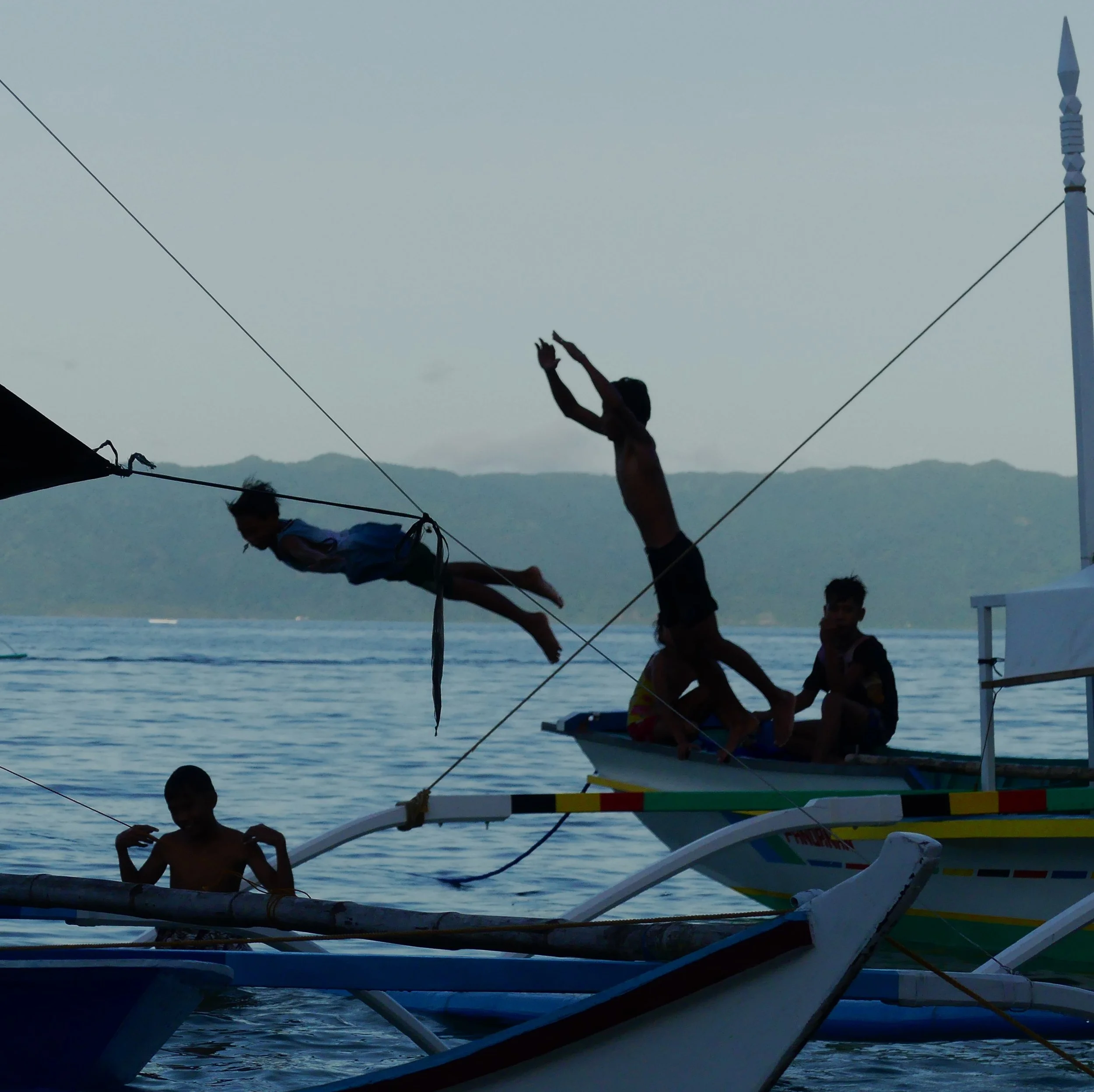 Children playing on boats and jumping into the water near the coast, with mountains in the background.