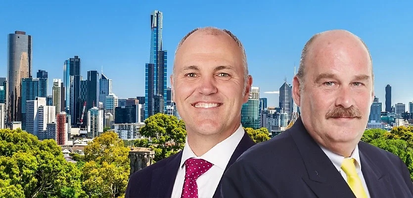 Two men in suits standing outdoors in front of a cityscape with tall skyscrapers and green trees.
