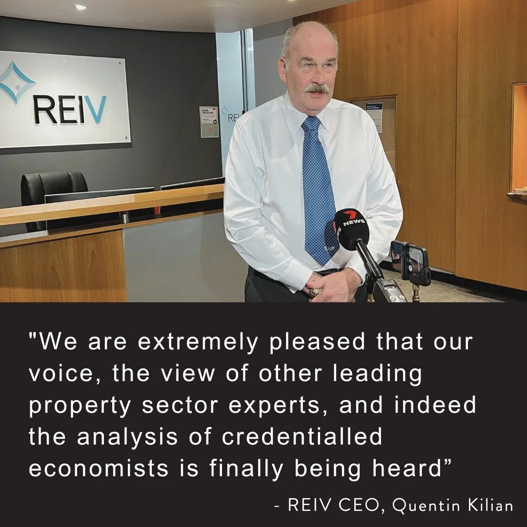 A man in a white shirt and blue tie speaking at a microphone during a press conference, with the REIV logo visible in the background.