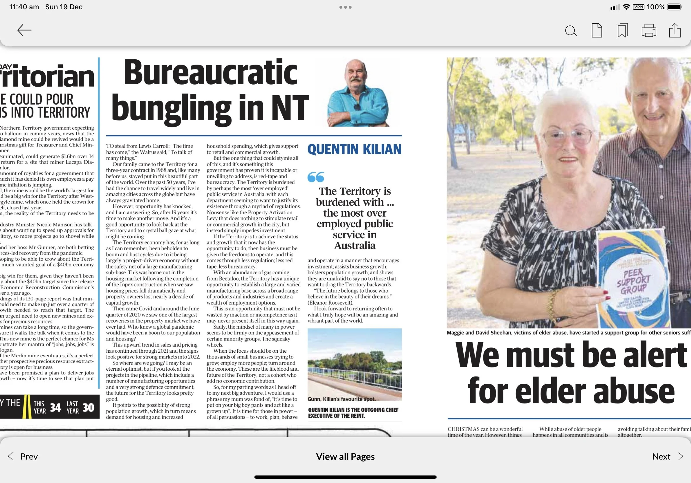 A woman and a man, both elderly, standing outdoors near a water body, holding a circular sign that reads 'Peer Support Group.' The woman has white hair, glasses, and is wearing a black and white patterned blouse, while the man has short gray hair, and is wearing a checkered shirt. They appear to be smiling. Trees with green leaves are visible in the background.