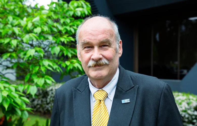 An older man with a mustache in a suit and yellow striped tie standing outdoors in front of green foliage.