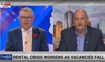Split-screen news broadcast showing two men in suits discussing a rental crisis, with a cityscape background and a news headline about worsening rental crisis as vacancies fall.