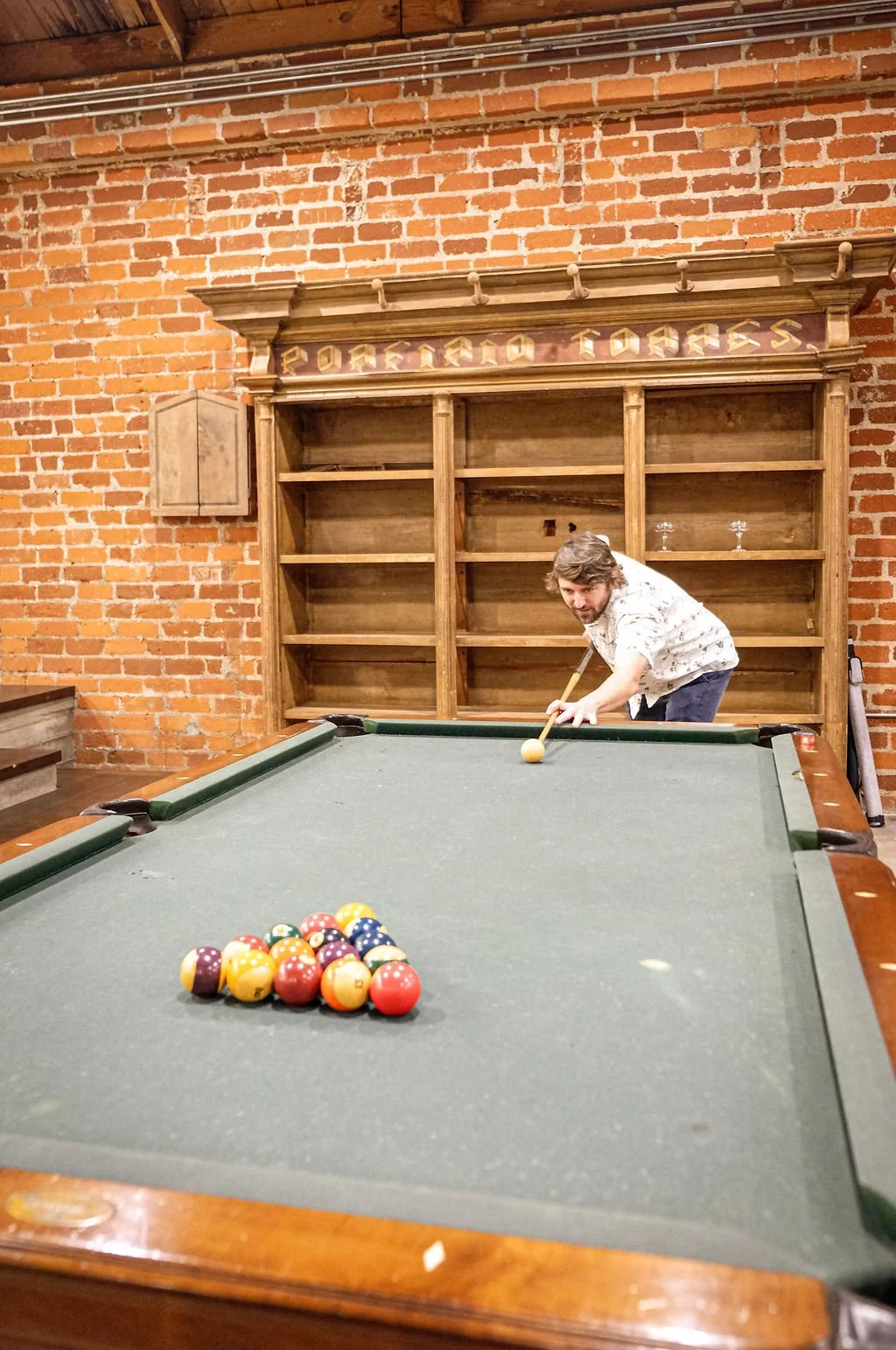 A man preparing to take a shot on a pool table with a brick wall and wooden shelves behind him.