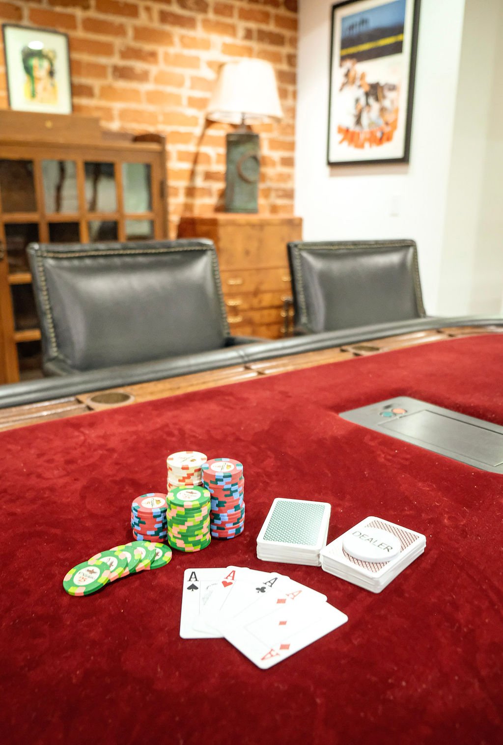 Poker game with chips, aces, and a deck of cards on a red felt table in a room with brick wall and framed posters.