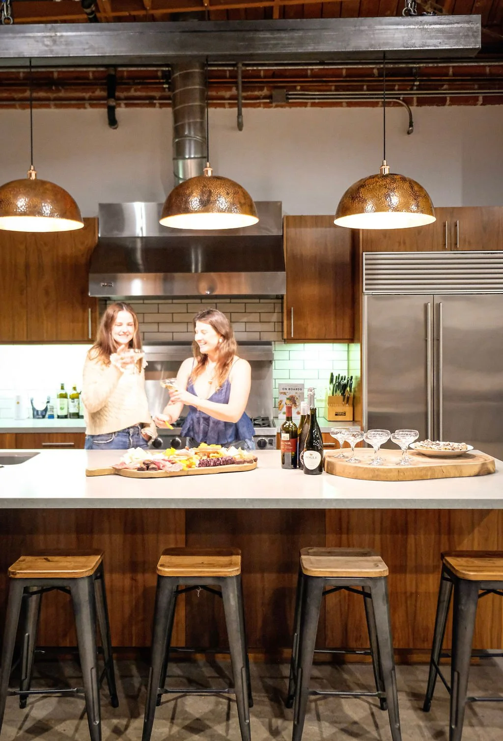 Two women standing in a modern kitchen, smiling and holding drinks, with charcuterie on a wooden platter, bottles of wine, glasses, and snacks on the island counters.