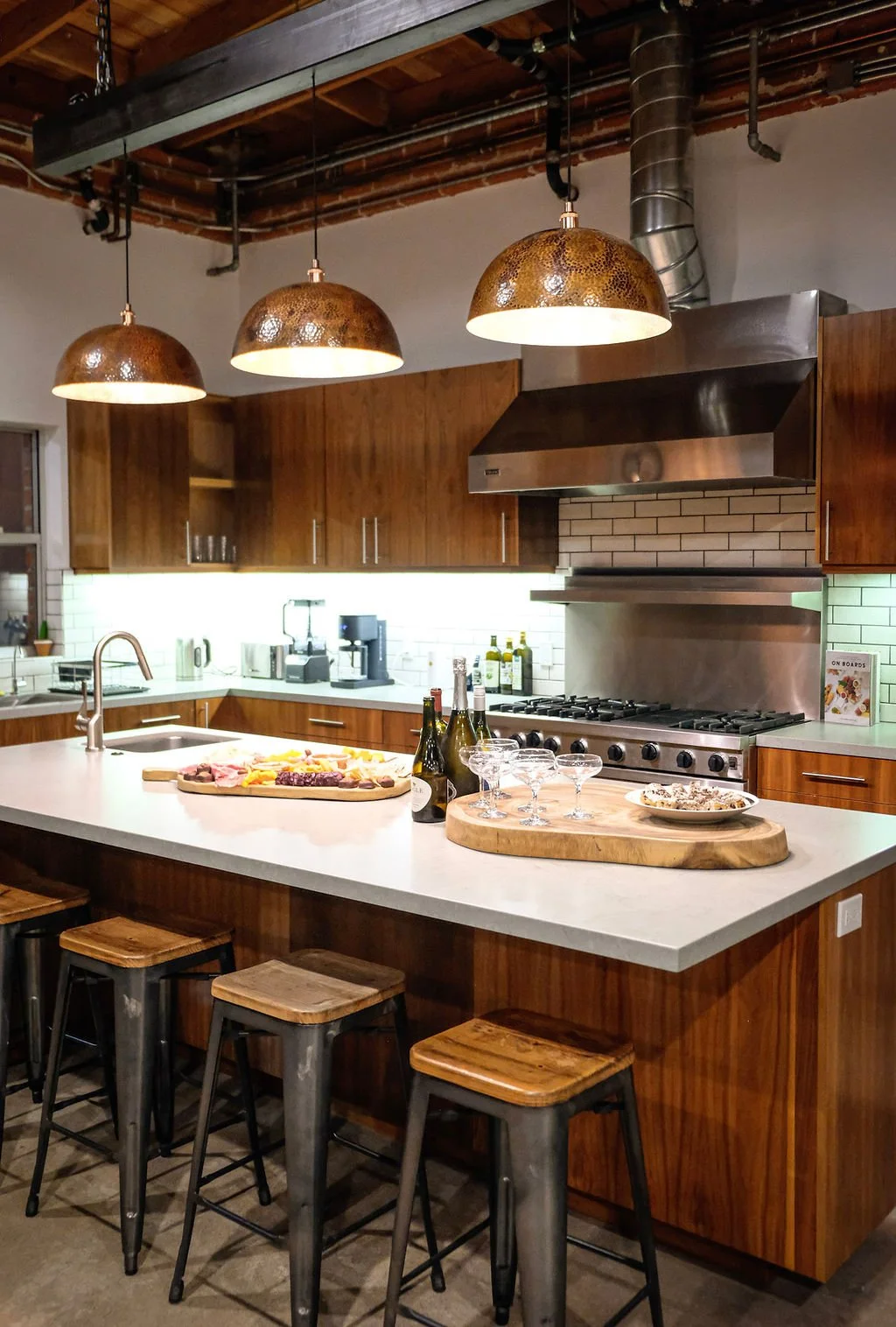 Modern kitchen with wooden cabinets, a white island counter with barstools, pendant lights, stainless steel appliances, and a brick backsplash. Items on the counter include an array of beverages, snacks, and glasses.