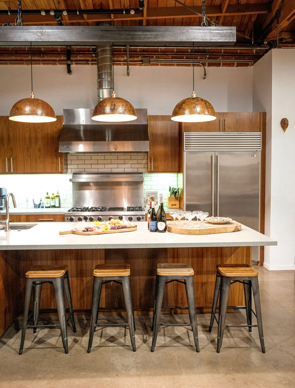 Modern kitchen with four wooden bar stools at an island, copper pendant lights, stainless steel appliances, and a countertop with bottles, glasses, and snacks.