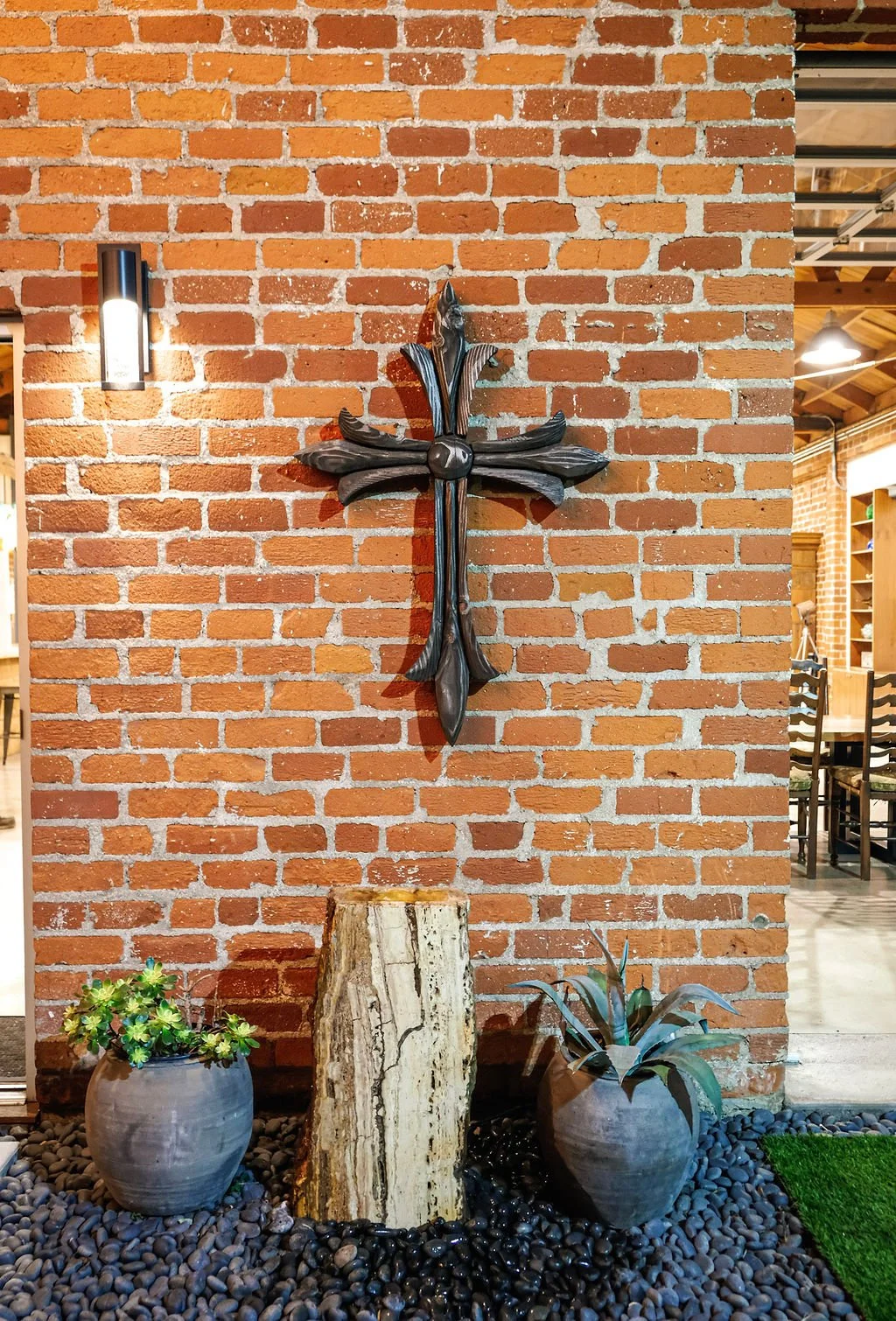 Decorative wall with a metal cross, two potted plants, and a vertical wood sculpture on a bed of black pebbles against a red brick wall.