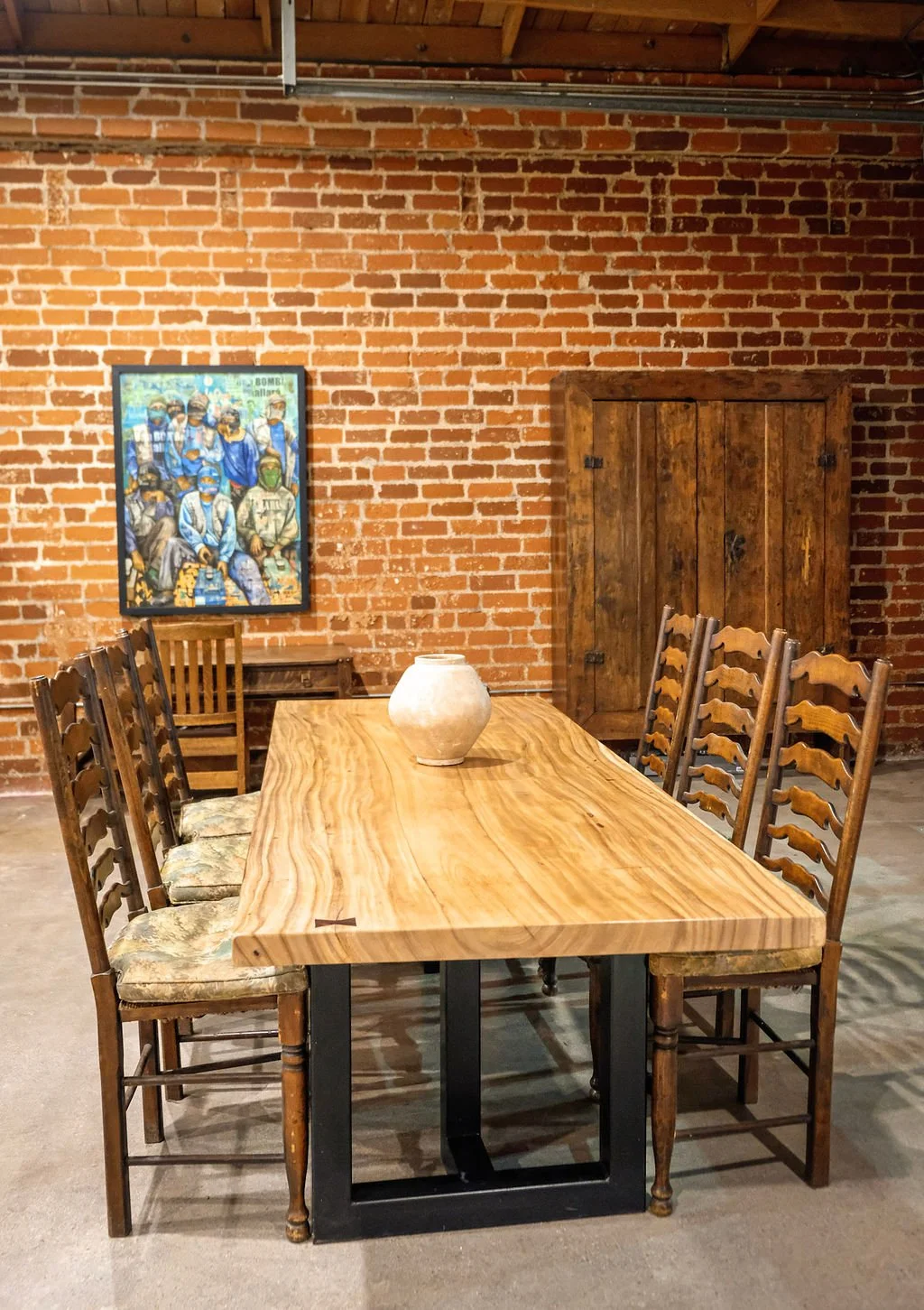 A rustic dining room with a wooden table and eight matching chairs, a white vase on the table, brick wall with a colorful painting, and a wooden cabinet against the wall.