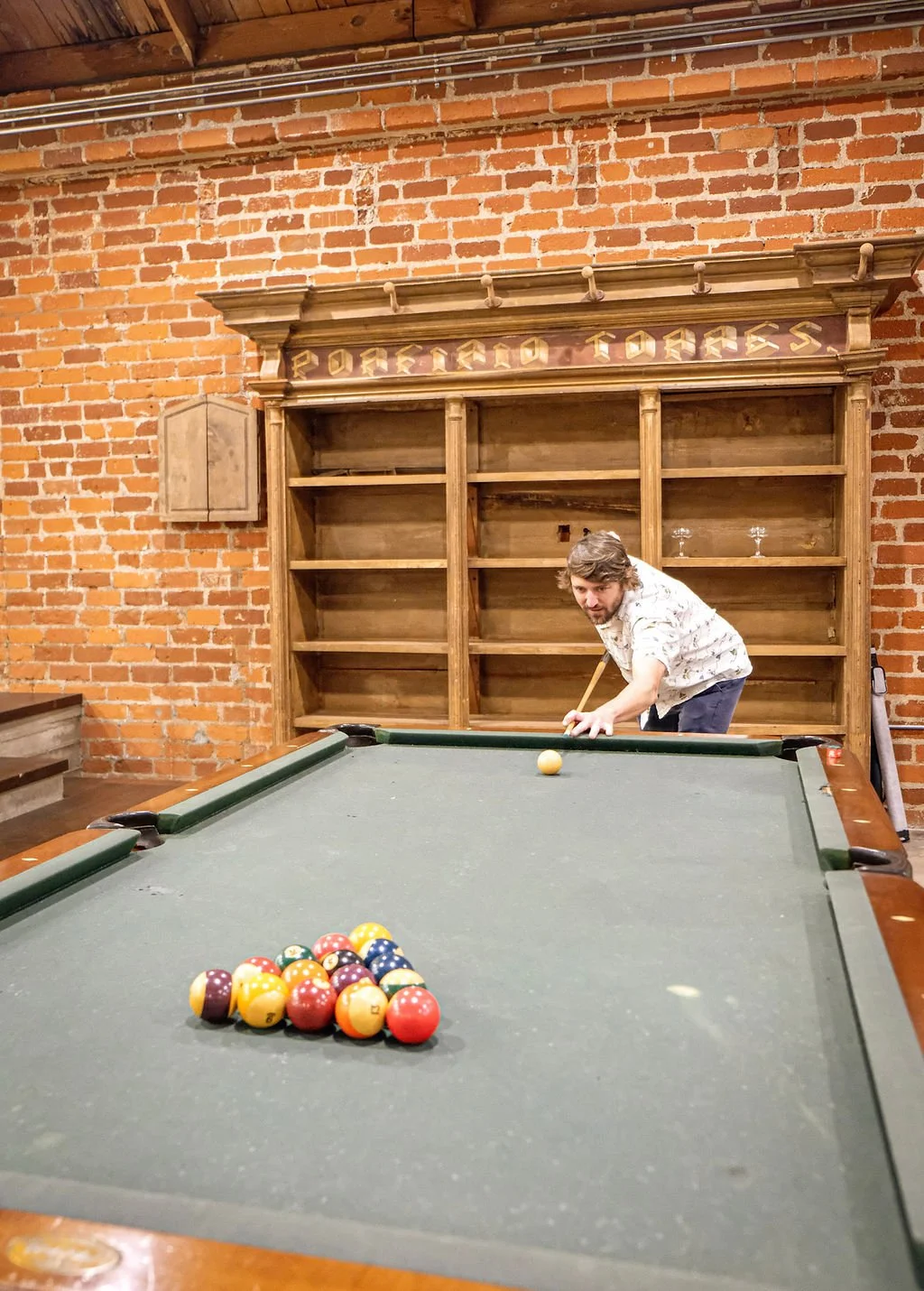 A man prepares to take a shot on a pool table in a bar with a brick wall and wooden shelves in the background.