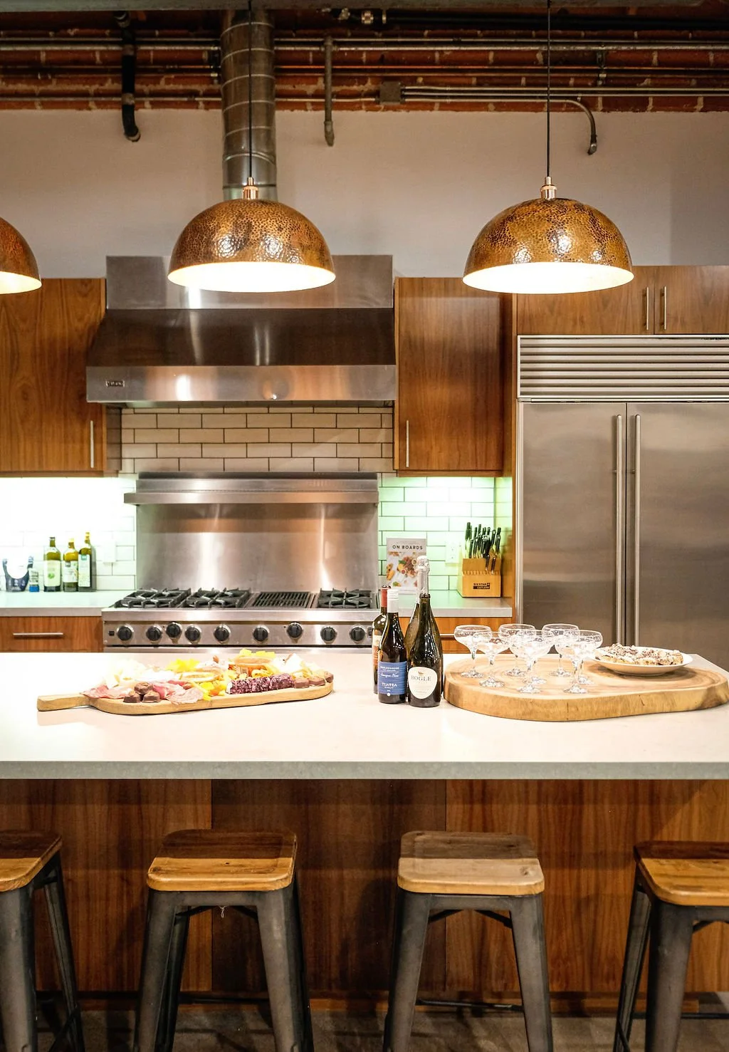Modern kitchen featuring pendant lights, a stainless steel range hood above a stove, wooden cabinets, a large refrigerator, a white countertop with drinks and appetizers, and bar stools in the foreground.
