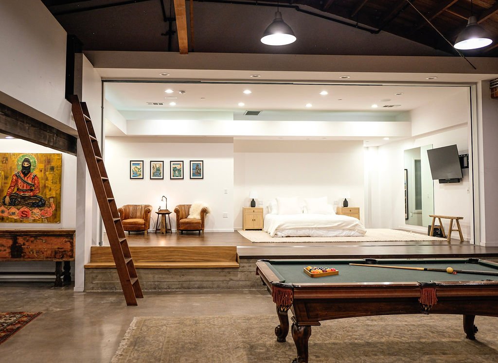 A spacious living area with a bedroom in the background, featuring a bed with white bedding, nightstands, and a small table. Two armchairs with a small table between them are on the left, and a large flat-screen TV is mounted on the wall on the right. There is a pool table in the foreground and artwork on the walls, with warm lighting and a loft-style industrial ceiling.