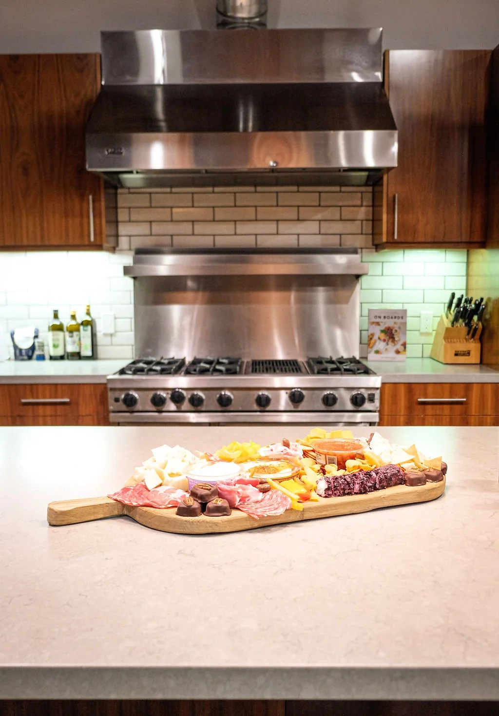 A wooden serving board with an assortment of cheeses, meats, chocolates, and condiments on a kitchen counter with a stove and wooden cabinets in the background.