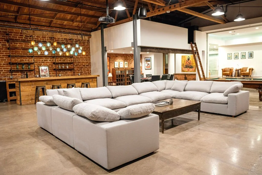 Spacious living area with a large white sectional sofa, wooden coffee table, exposed brick wall, modern light fixtures, and a bar area with stools in an industrial-style space.