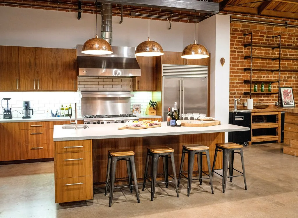 A modern kitchen with wooden cabinets, a large island with four bar stools, and stainless steel appliances. The kitchen features a brick wall and open shelving on the right side.