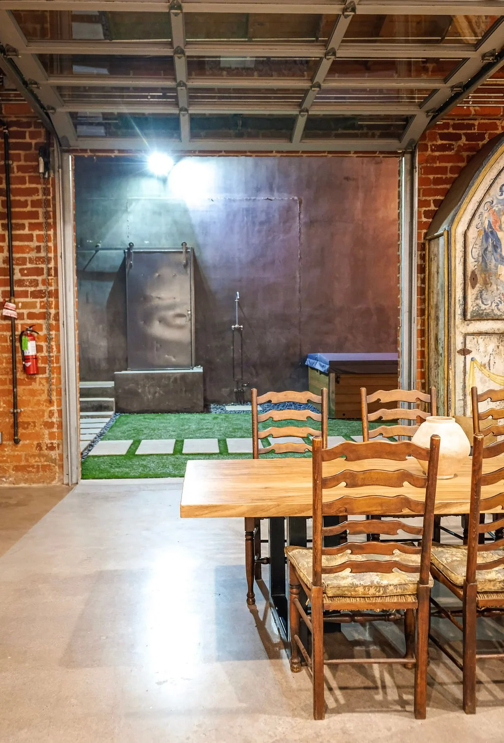 A dining area with a wooden table and six chairs, a decorative ceramic vase on the table, brick walls, and a rustic shower area outside.