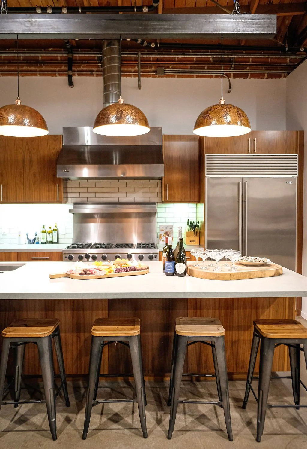 Modern kitchen with a white countertop island, four metal and wood barstools, stainless steel appliances, wooden cabinets, pendant lights, wine bottles, glasses, and a cheese platter.