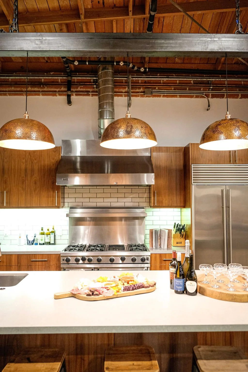 Kitchen with a white island counter, wooden cabinets, stainless steel appliances, and hanging pendant lights. On the counter, there is a cheese and charcuterie board, bottles of wine, and glasses.
