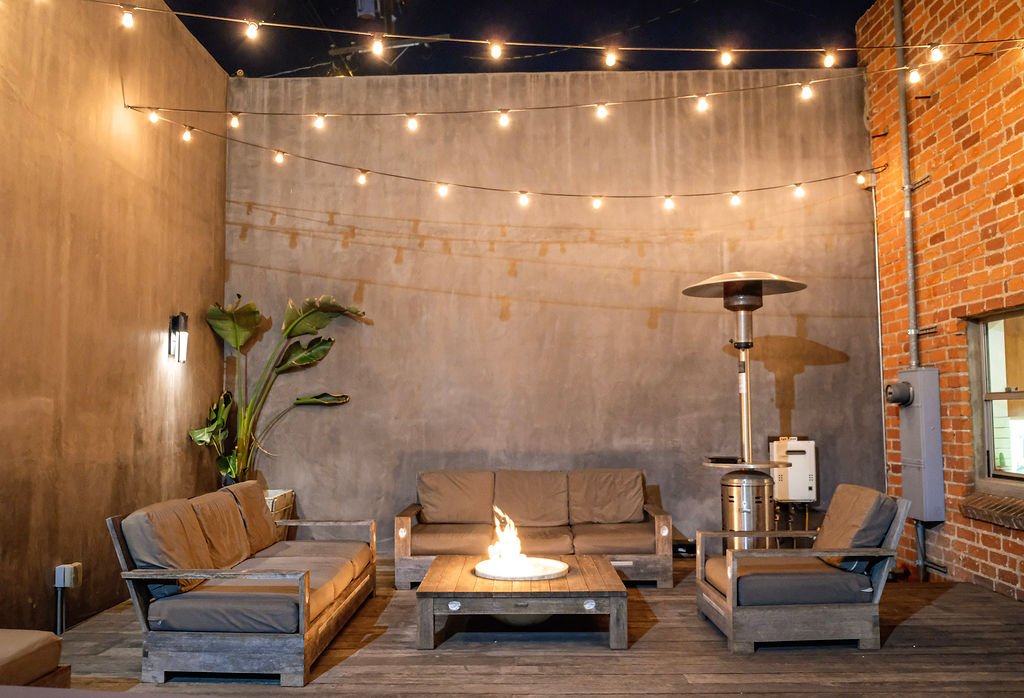 Outdoor patio with string lights, surrounded by brick and concrete walls, furnished with wooden sofas, a fire pit table, a patio heater, and potted plants.