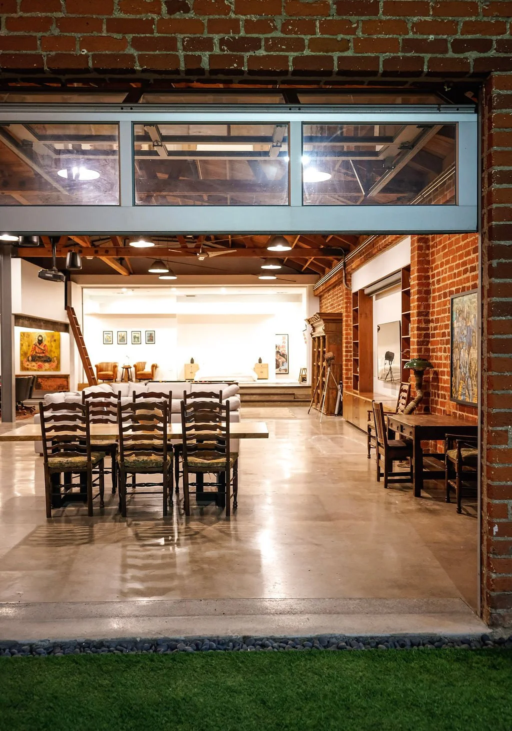 Interior of a spacious room with brick walls, wooden beams, and various chairs, a sofa, and artwork, possibly a gallery or event space.