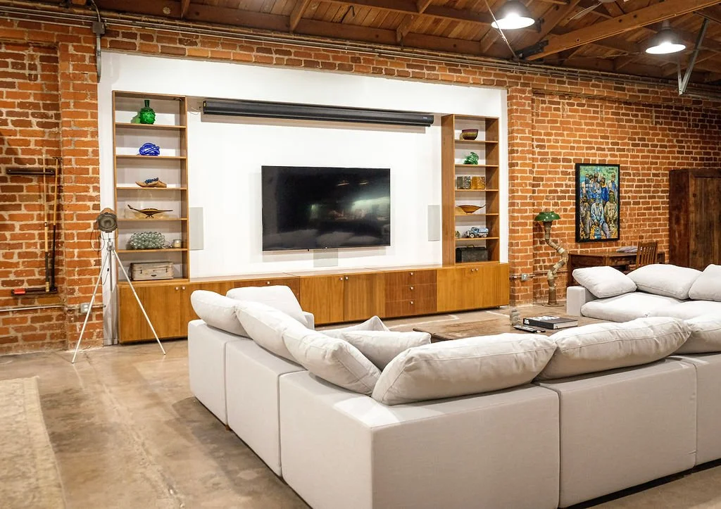 Living room with exposed brick walls and ceiling, a large white sectional sofa with multiple cushions, a mounted flat-screen TV on a white wall section, wooden shelves with decorative items, a vintage tripod floor lamp, a framed artwork, and a small wooden desk with a chair.