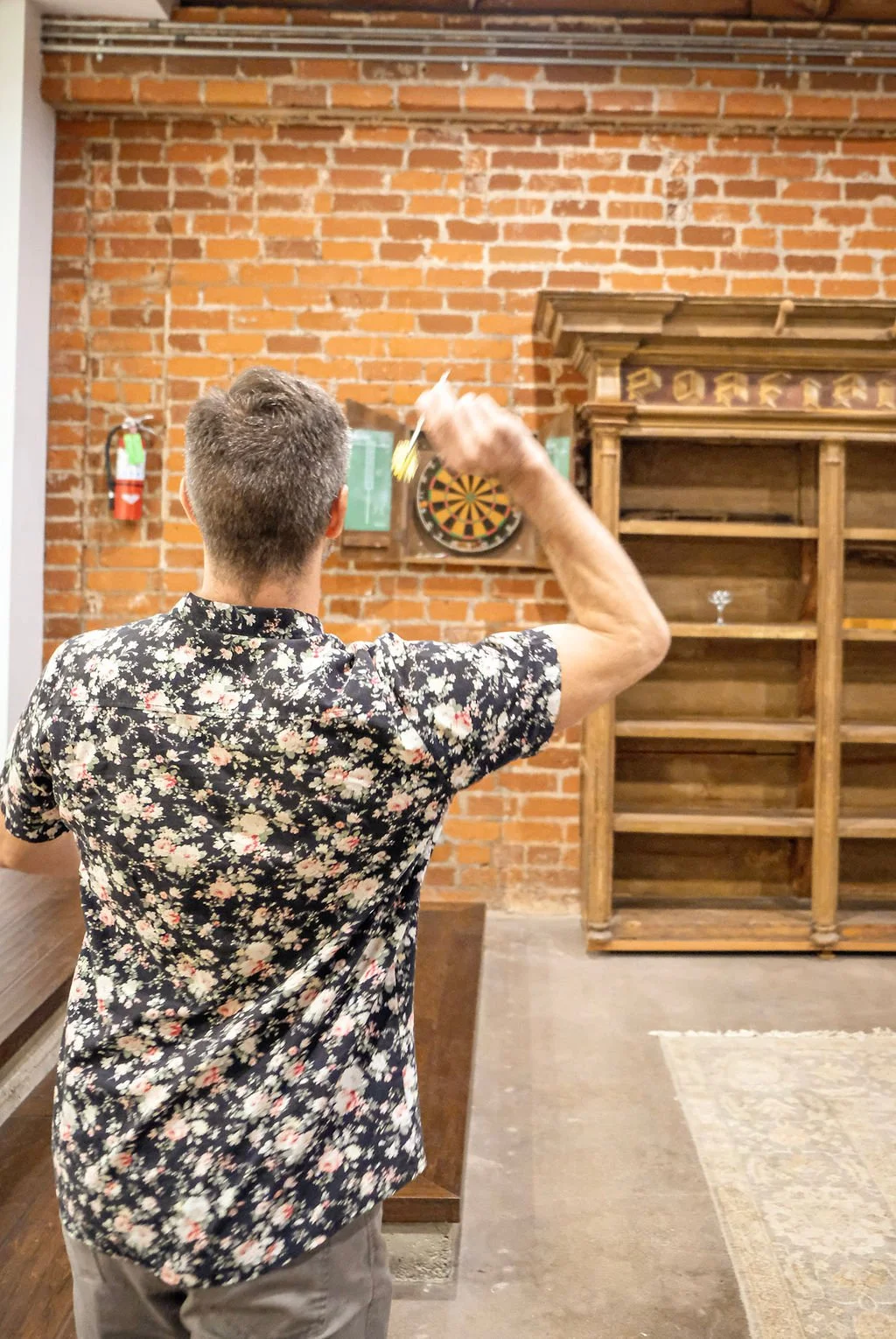 A man with short hair wearing a black floral shirt is playing darts in a room with brick walls and wooden furniture.