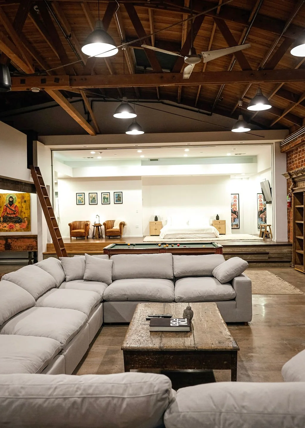 A spacious living area with a large white sectional sofa, a rustic wooden coffee table, and an open bedroom space in the background with a bed, side tables, and chairs. The ceiling features wooden beams, a ceiling fan, and industrial-style lights.