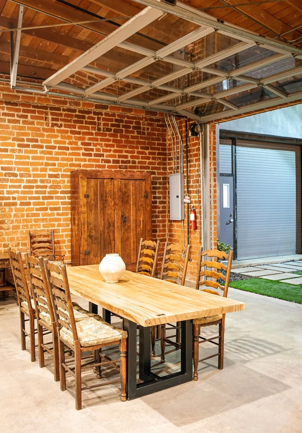 Wooden dining table with eight chairs and a white vase, industrial-style exposed brick wall with a wooden door, garage with metal rolling door, and open garage door showing outdoor space with greenery.