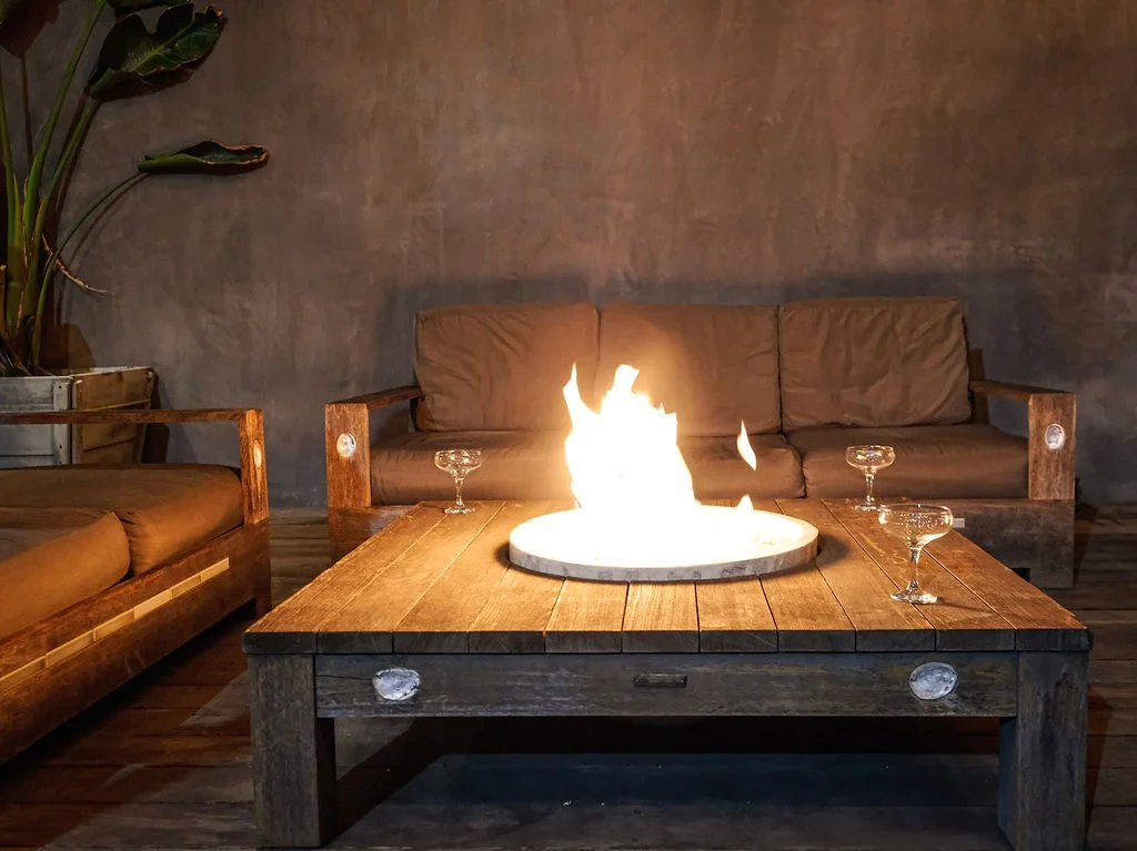 Living room with a wood coffee table and a fire pit, surrounded by sofas and glasses, beige sofas, potted plant in the corner, gray wall, wooden floor.