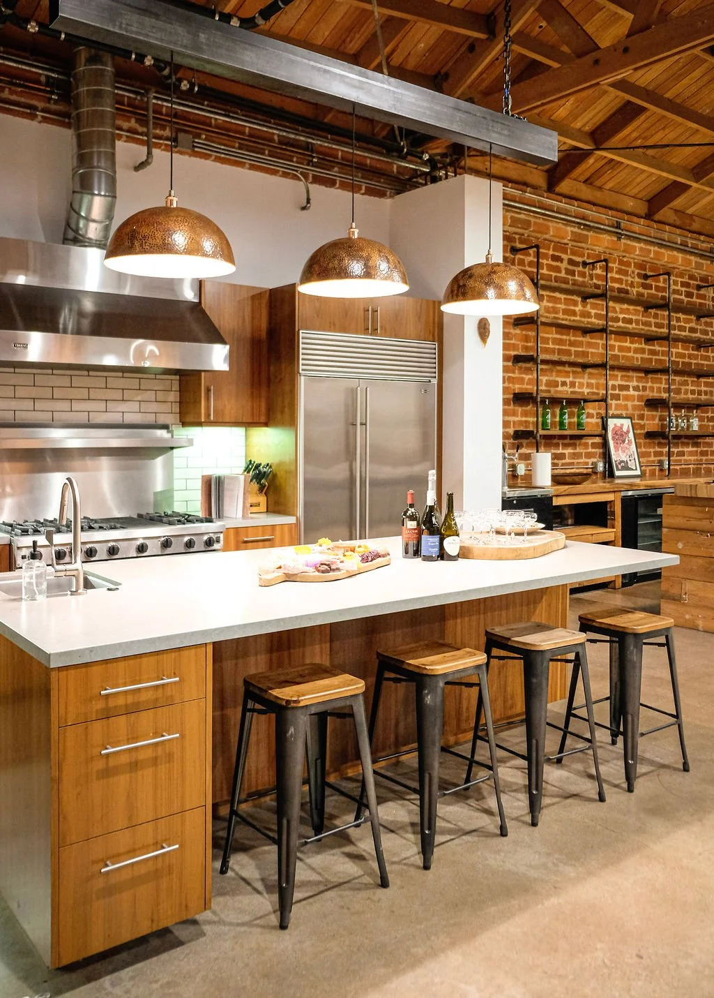 Modern industrial kitchen with wooden cabinets, stainless steel appliances, a white kitchen island with bar stools, and three copper pendant lights hanging above.