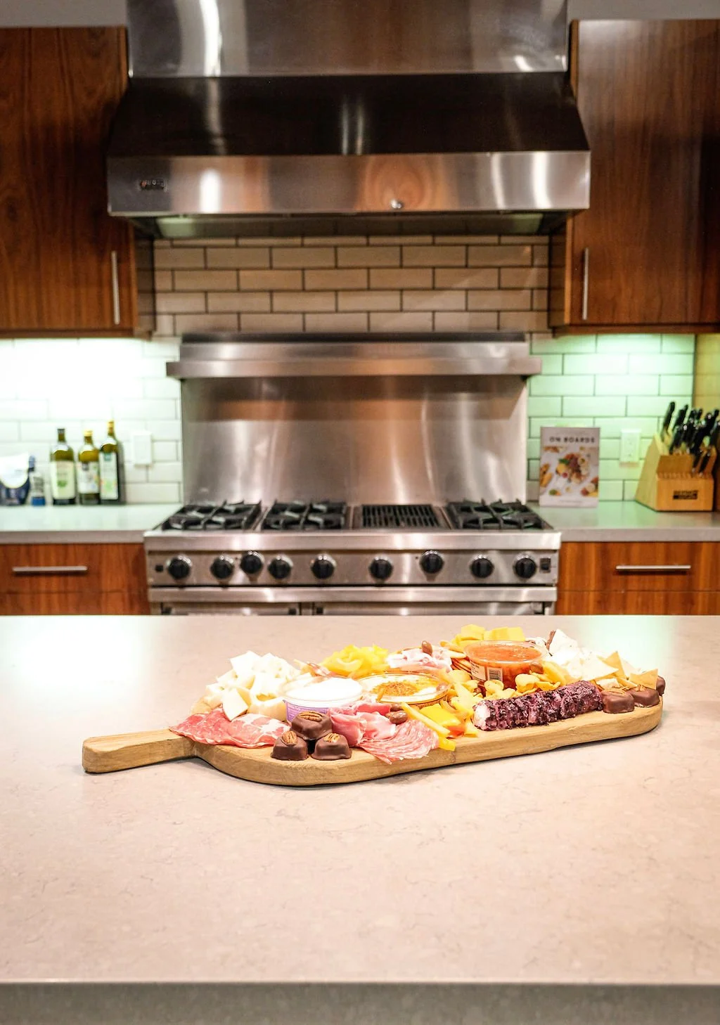 A charcuterie board with assorted meats, cheeses, and condiments on a kitchen countertop, with a professional stove and green backsplash in the background.