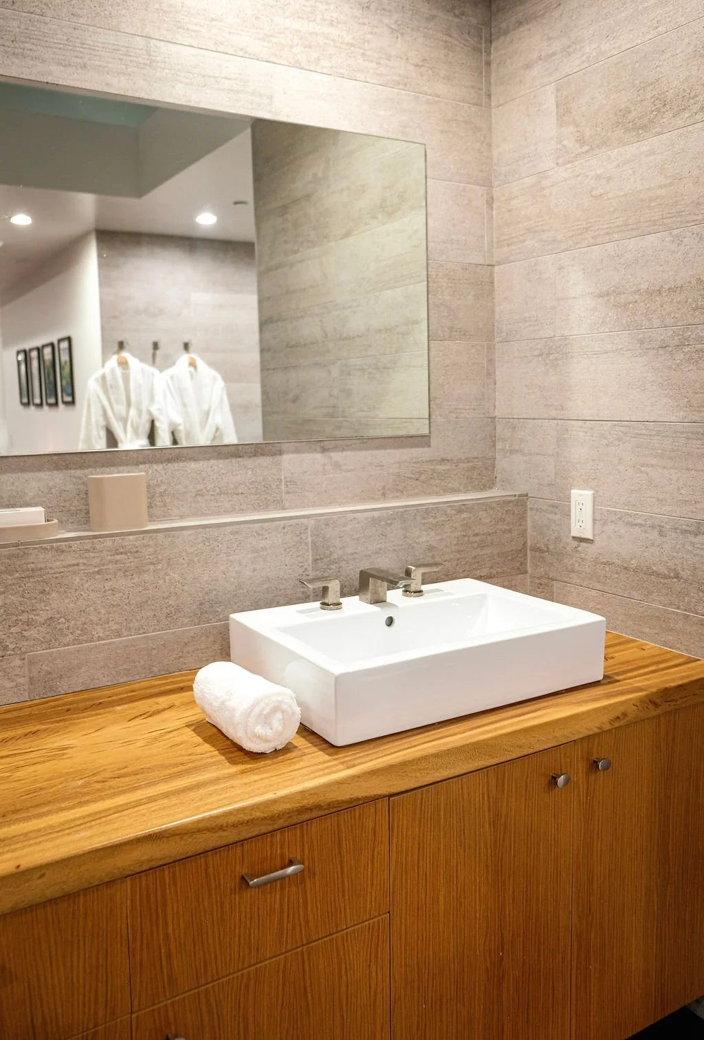 Modern bathroom sink with a rolled white towel on a wooden countertop, large mirror, beige tiled walls, soap dispenser, and electrical outlet.