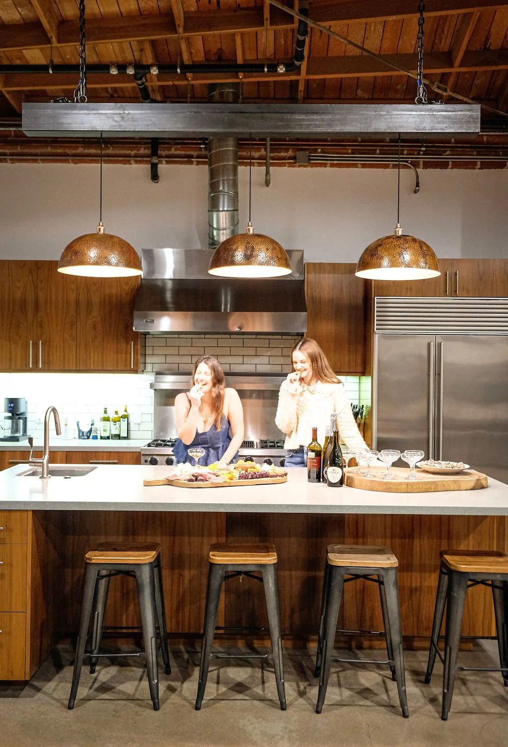 Two women laughing and eating at a kitchen island with drinks and snacks, in a modern kitchen with wooden cabinets and industrial-style pendant lighting.