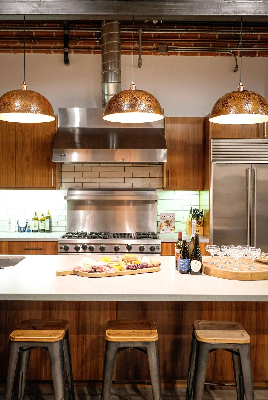 Kitchen with three hanging copper pendant lights, a stainless steel range hood, a stove, wooden cabinets, a refrigerator, a cutting board with cheese and meats, bottles of wine, and empty wine glasses on a round wooden tray.