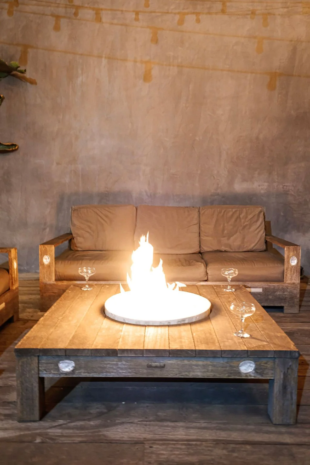 A cozy living room with a beige couch, a wooden coffee table with a fire pit, and four empty glass cups on the table. There is a neutral-toned textured wall in the background and a plant partially visible on the left.