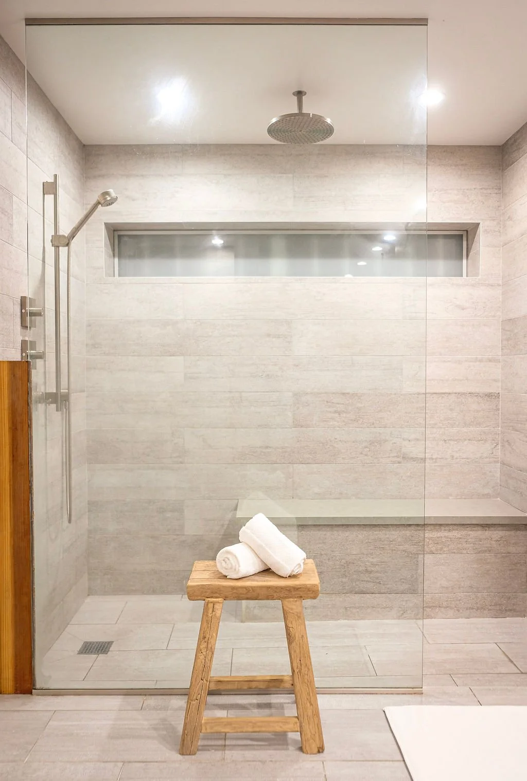 Walk-in shower with beige tiles, a wooden stool with rolled white towels, a square shower drain, a horizontal window, and a ceiling-mounted shower head.