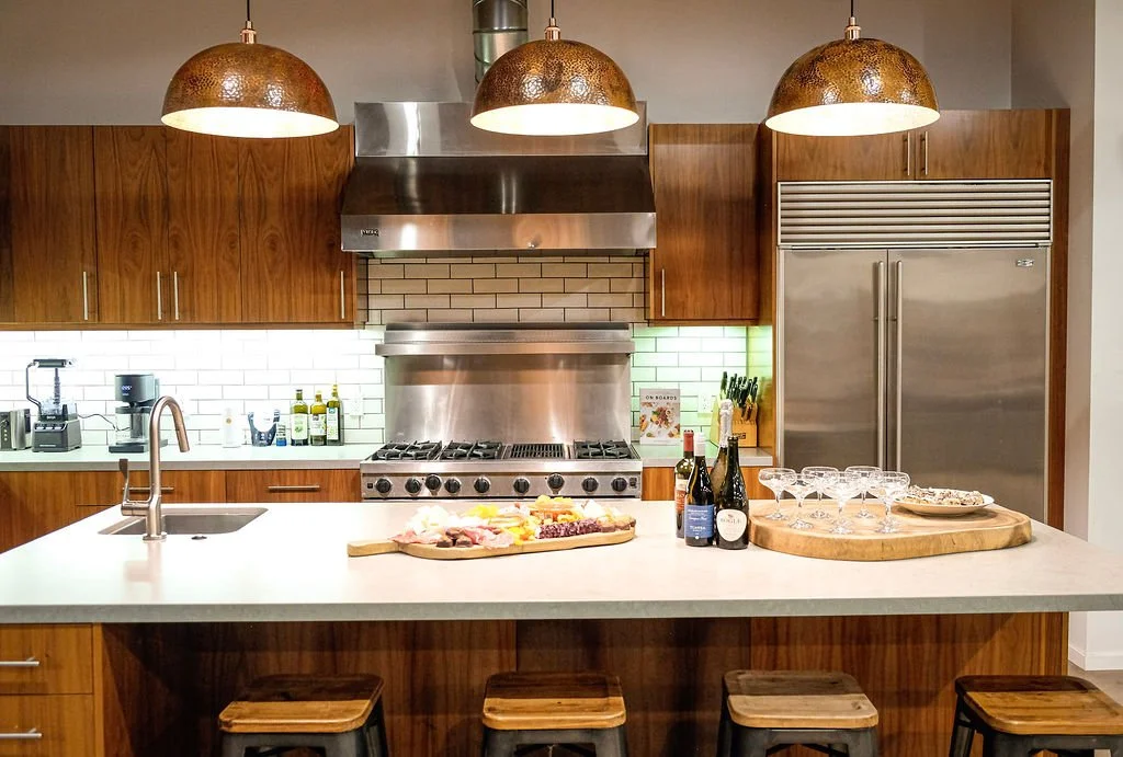 Modern kitchen with wooden cabinets, stainless steel appliances, and pendant lights. Countertop with wine bottles, glasses, cheese platter, and a wrapped meat and cheese board.