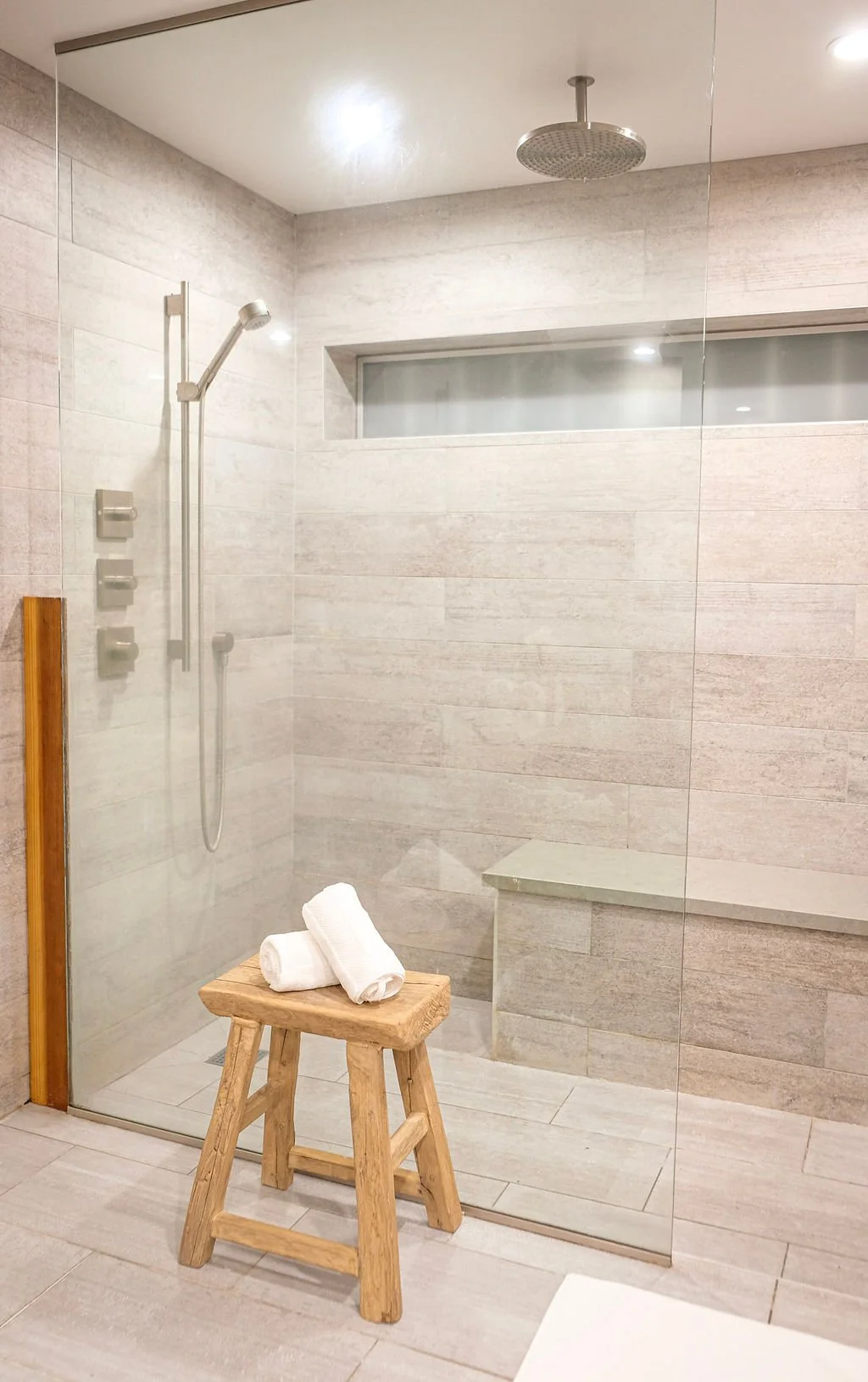 Modern walk-in shower with a glass enclosure, rain shower head, and wall-mounted shower controls, with a wooden stool holding rolled white towels.