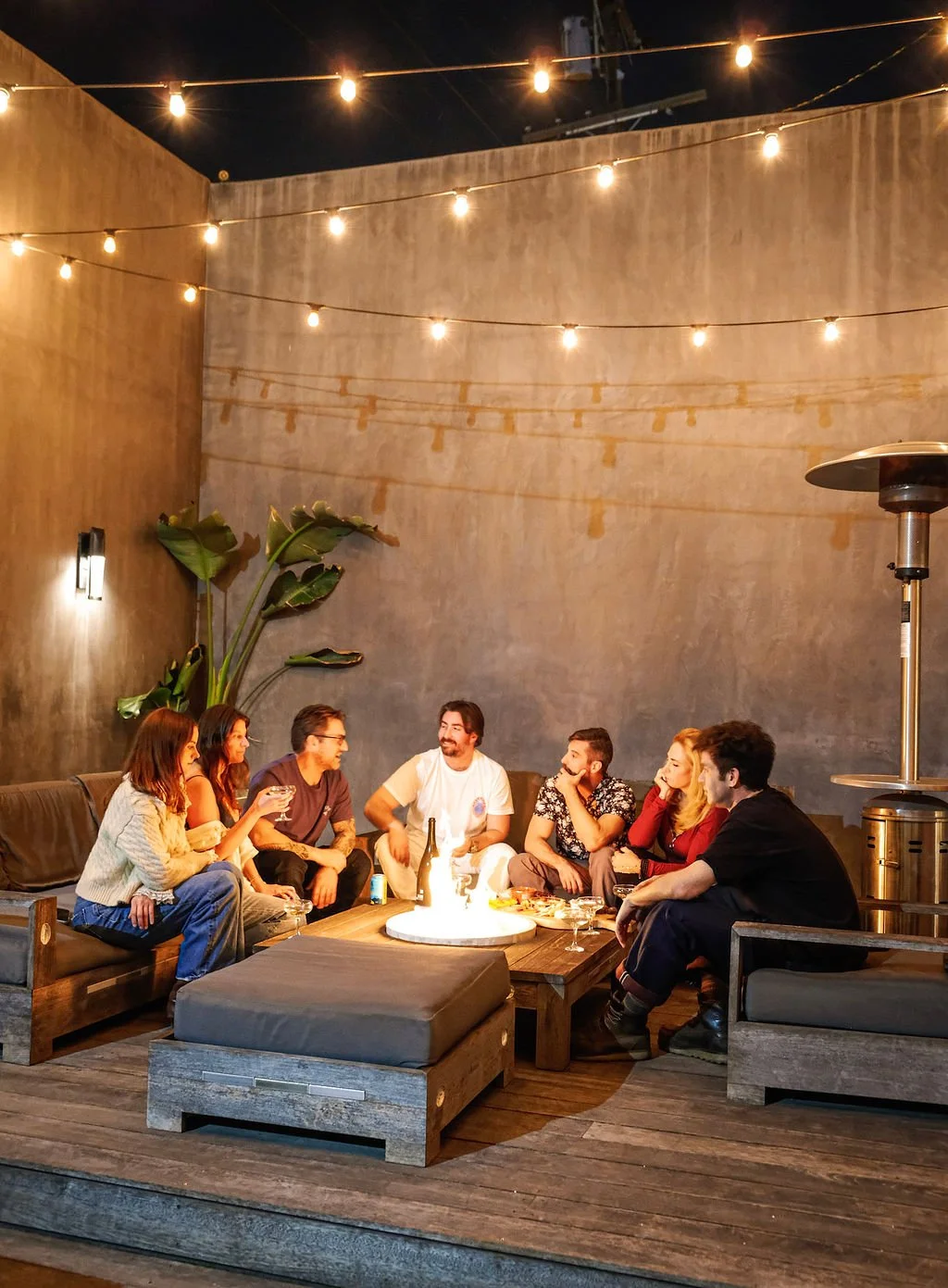 A group of seven young adults sitting on a wooden patio with string lights overhead, enjoying drinks and conversation around a low table with snacks, in an outdoor space with concrete walls and large leafy plants.