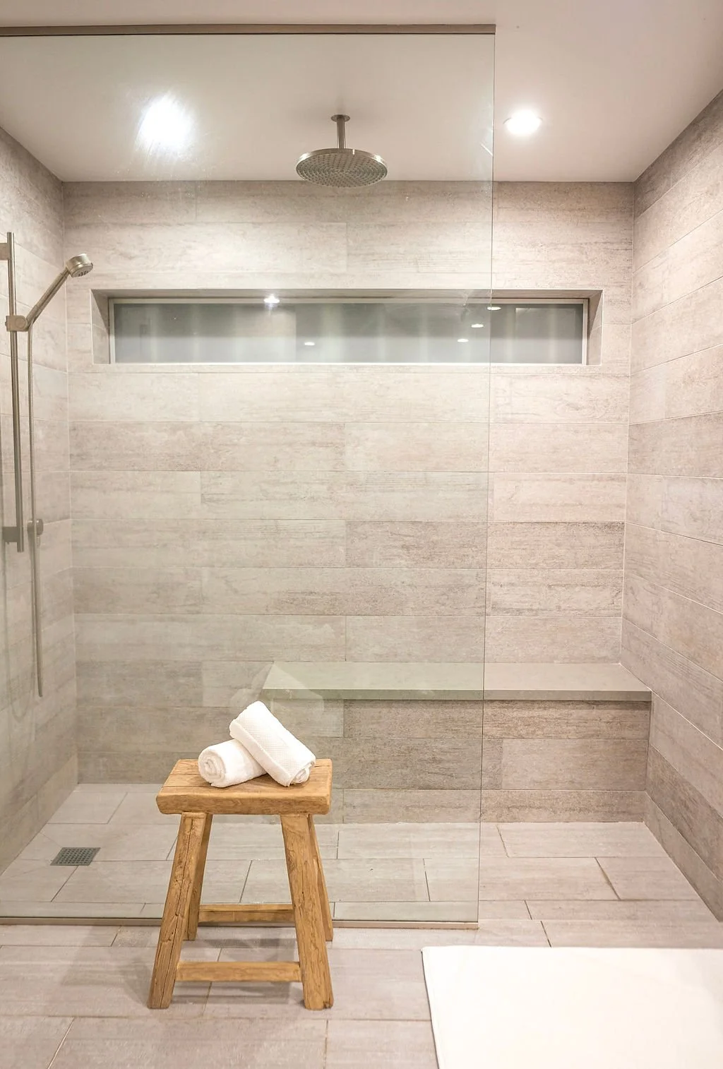 Modern walk-in shower with gray tiled walls and floor, a ceiling-mounted rainfall showerhead, a small window near the ceiling, a wooden stool with rolled white towels, and a glass partition.