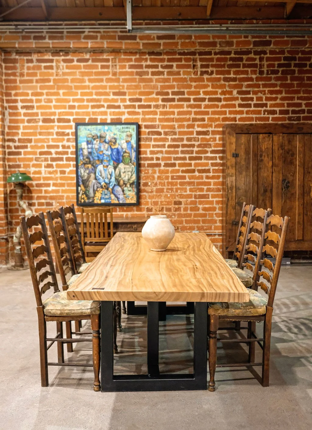 A rustic dining room with a long wooden table, nine chairs, a decorated ceramic vase, a red brick wall with a painting of group of people, a wooden cabinet, and a green desk lamp.