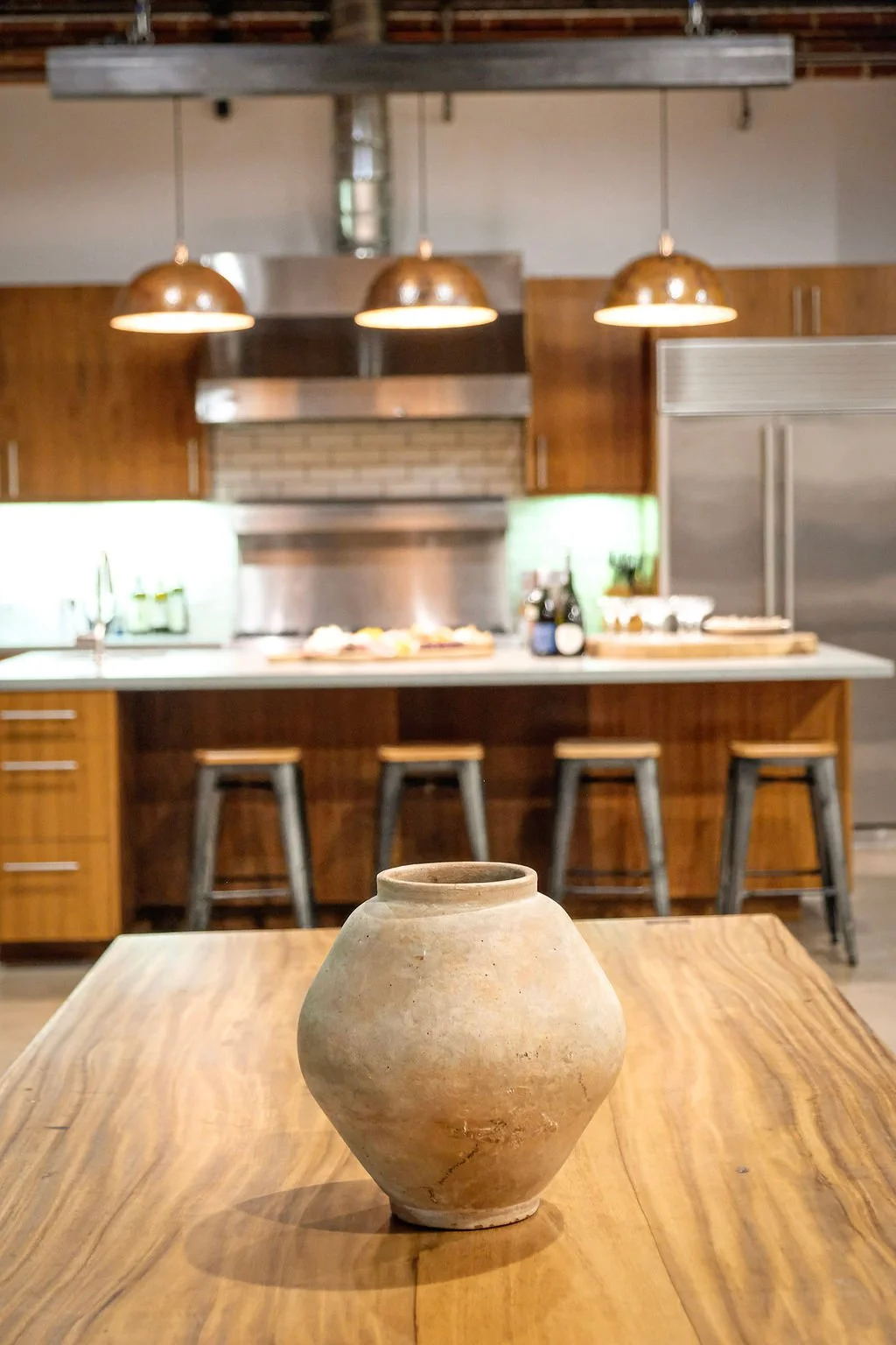 An earthenware vase on a wooden table with a rustic kitchen in the background featuring a countertop, bar stools, kitchen appliances, and hanging copper pendant lights.