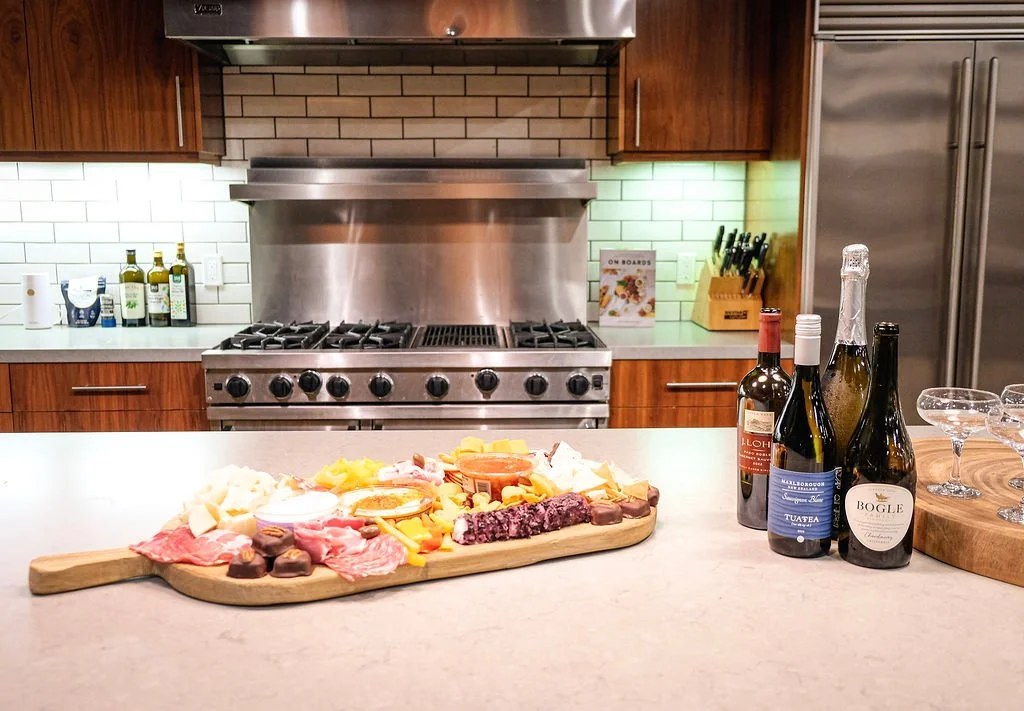 A kitchen with a countertop display of assorted cheeses, meats, and chocolates on a wooden platter, and four bottles of wine with three empty wine glasses nearby.