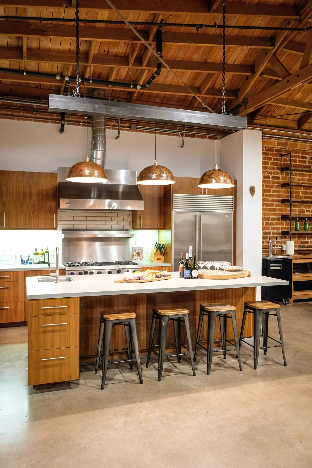 Kitchen with wooden cabinets, stainless steel appliances, and a white kitchen island with four stools. Hanging copper pendant lights above the island, exposed wooden ceiling with visible beams, and brick wall detail.