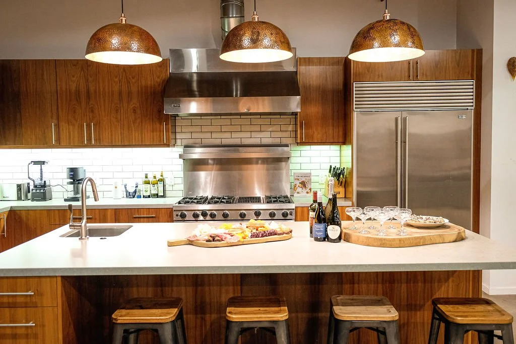 Modern kitchen with wooden cabinets, a white countertop island, stainless steel appliances, and hanging pendant lights. The island has a wooden tray with glasses and a charcuterie board with various foods, and a few bottles of wine.