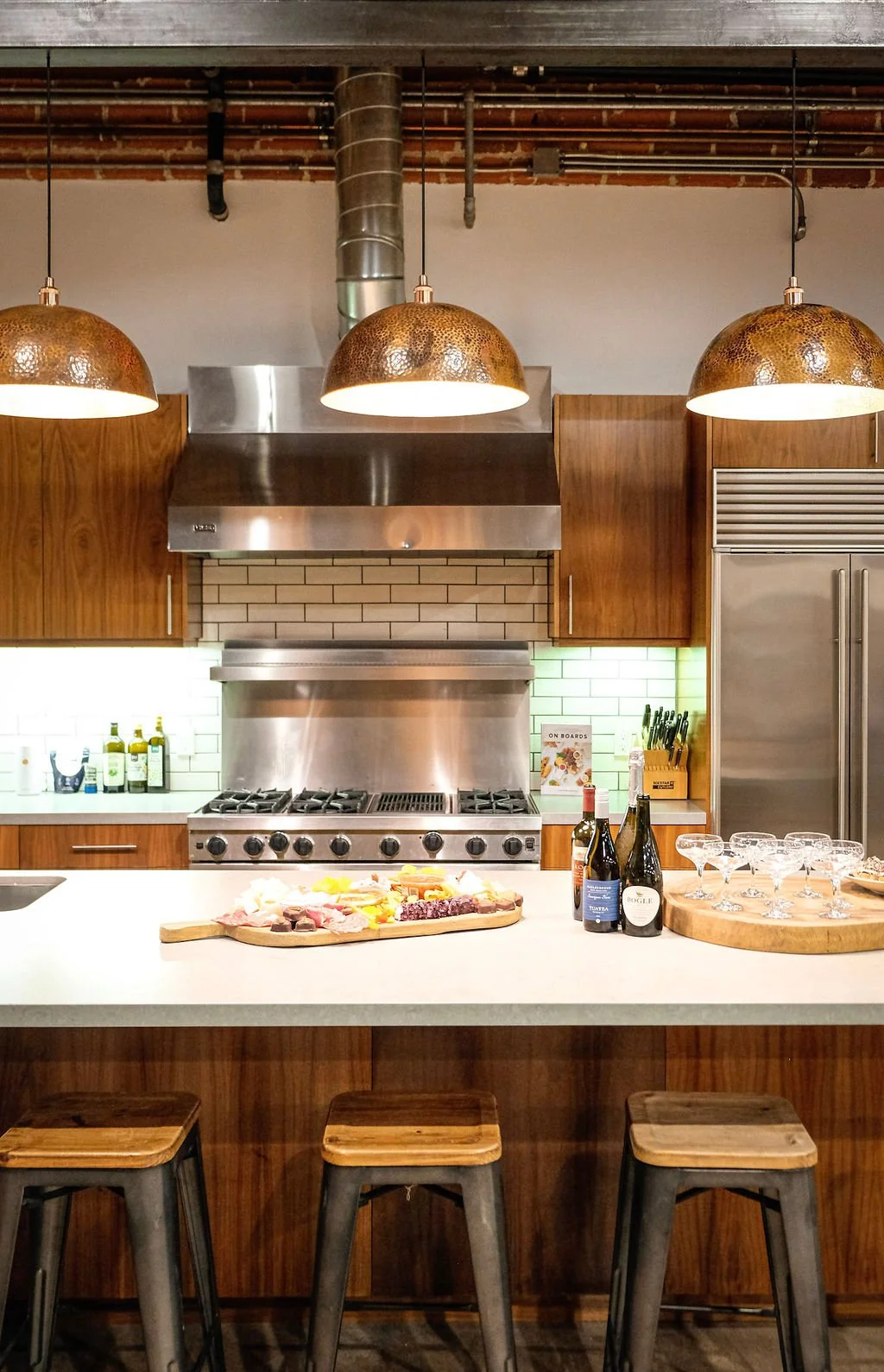 Modern kitchen with wooden cabinets, stainless steel appliances, hanging copper pendant lights, a white countertop with a charcuterie board, bottles of wine, glasses, and three wooden and metal stools in the foreground.