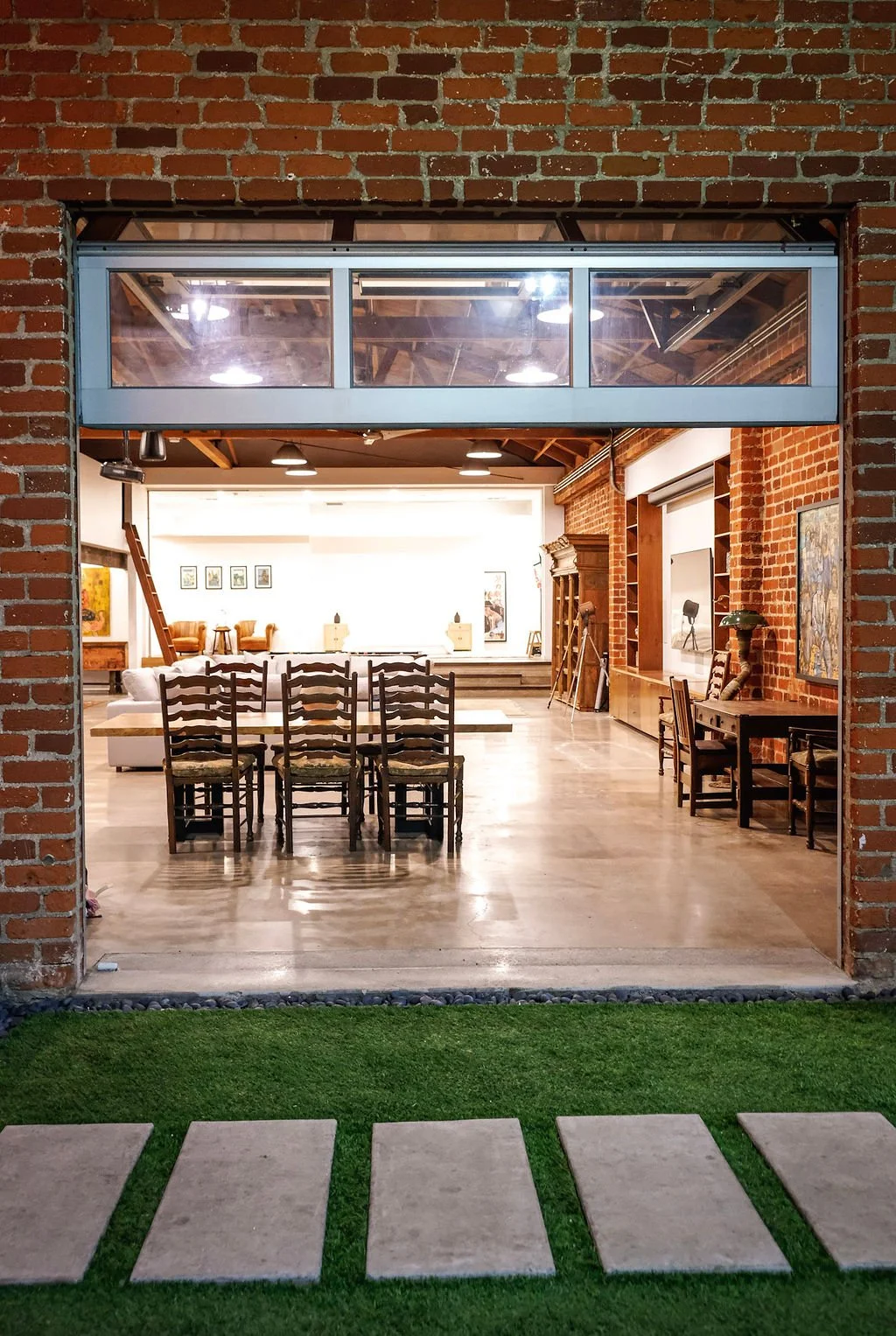 View from outside into an open-concept interior with red brick walls, wooden furniture including a long dining table with nine chairs, and a stage area with artwork and seating, illuminated by ceiling lights.