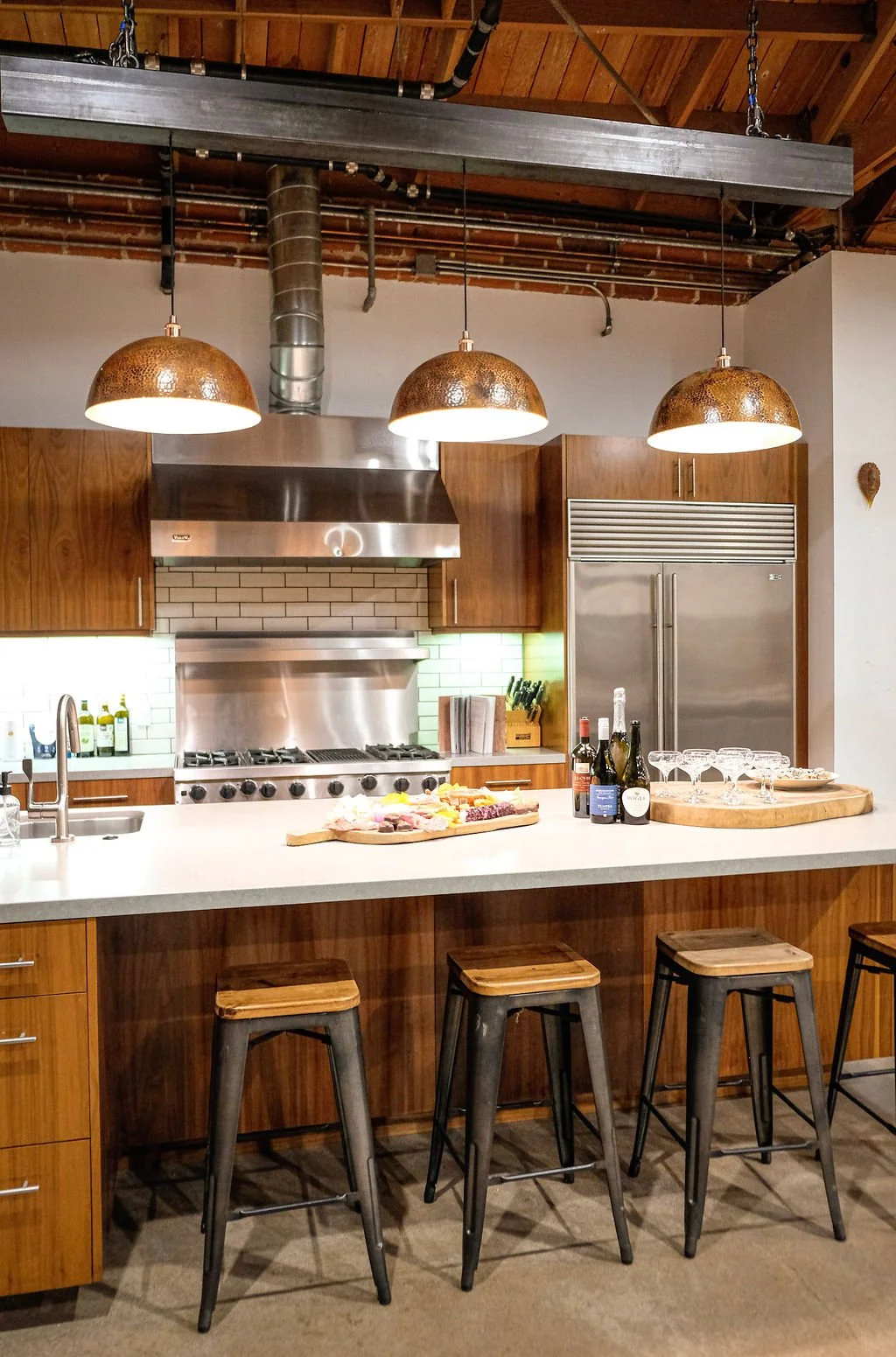 Modern kitchen with a white island counter, three metal and wood bar stools, stainless steel appliances, wooden cabinets, pendant lights, and a platter of food, wine bottles, and glasses on the counter.
