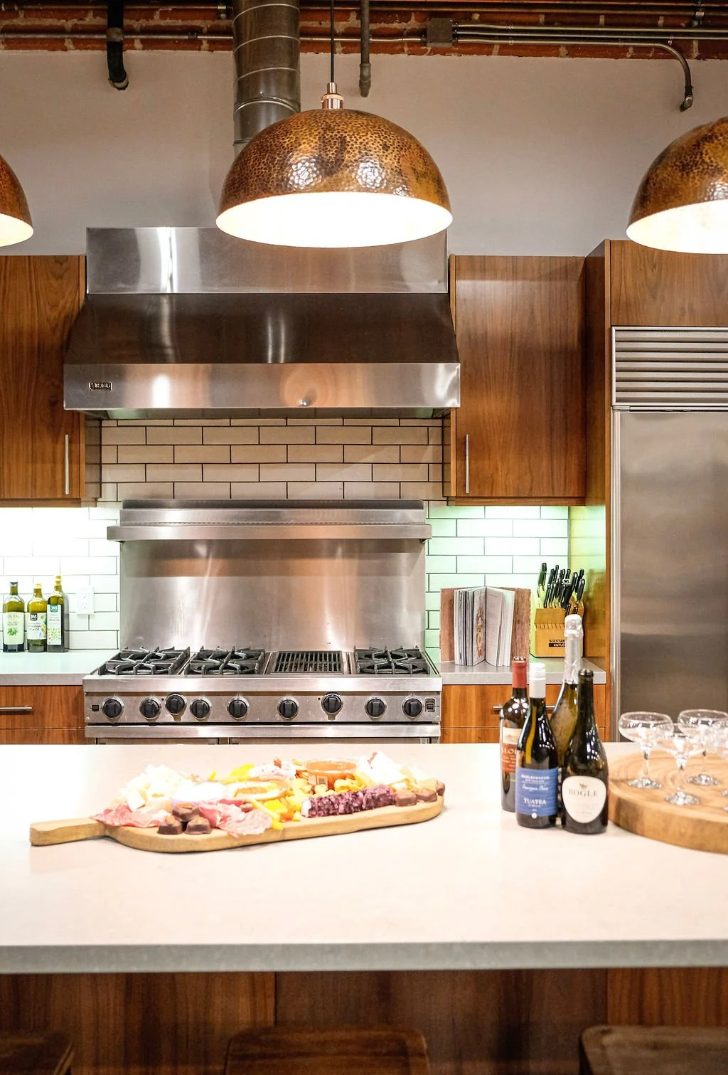 Kitchen with modern stainless steel stove, wooden cabinets, and pendant lights, with a cheese and charcuterie platter, wine bottles, and wine glasses on the white countertop.
