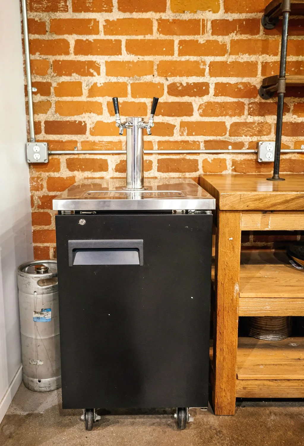 A beverage Kegerator with two taps, situated next to a wooden workbench on a brick wall background.